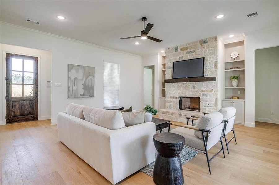 Living room featuring built in features, a stone fireplace, light hardwood / wood-style floors, ceiling fan, and ornamental molding