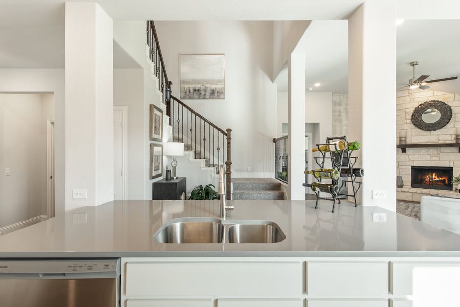 Kitchen island with double sink and gray countertop, open to living room with stone fireplace and staircase