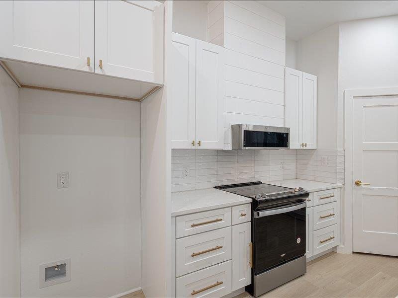 This kitchen features white cabinetry with gold hardware, a stainless steel microwave and stove, a light-colored tile backsplash, and light countertops