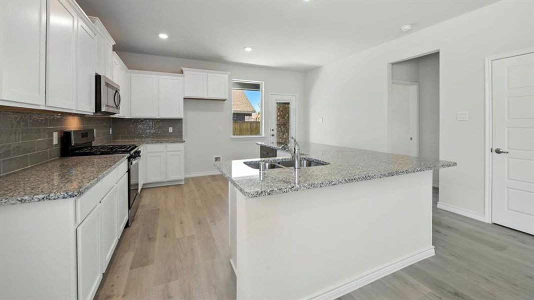 Kitchen featuring appliances with stainless steel finishes, a kitchen island with sink, light stone countertops, tasteful backsplash, and light wood-type flooring