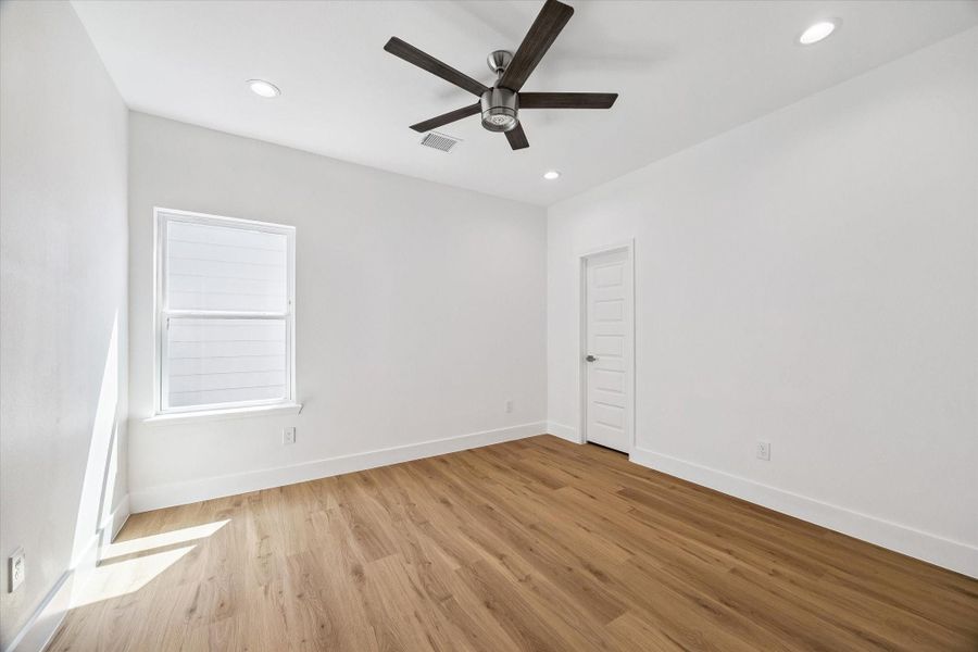 This bright secondary bedroom offers warm wood-look flooring, neutral walls, and a modern ceiling fan for a clean contemporary feel. A large window brings in natural light, while recessed lighting adds additional illumination. The simple, spacious layout provides flexibility for bedroom furnishings, a home office arrangement, or a comfortable guest retreat.