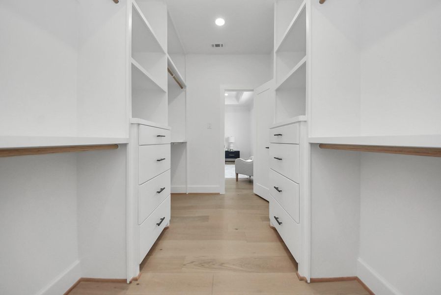 Step into this expansive walk-in closet, featuring ample shelving, drawers, and hanging space. Light wood flooring and white cabinetry create a bright, modern environment, offering plenty of storage and organization for everyday living.