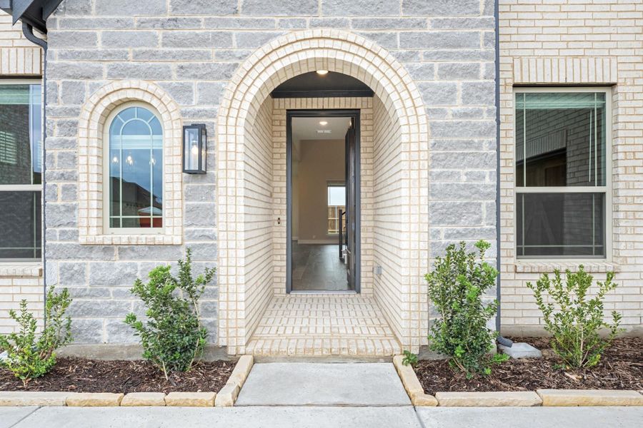 Home entrance with arched brick surround, open front door, stone exterior, and small shrubs on each side