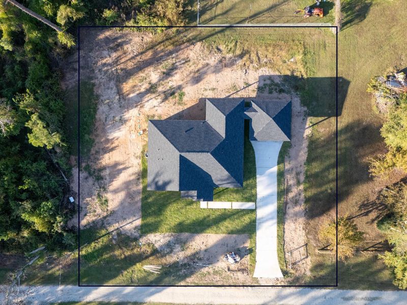 This aerial photo shows a newly constructed home with a dark roof, situated on a spacious lot surrounded by trees. The property features a long driveway leading to the garage, a neatly maintained lawn, and a partially wooded area providing privacy.