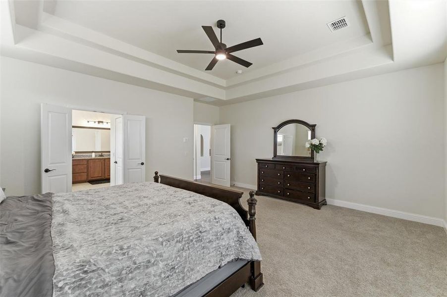Bedroom with light colored carpet, a ceiling fan, a raised ceiling, and ensuite bath