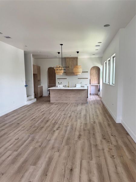 This photo showcases a spacious, modern kitchen with an open floor plan. It features light wood flooring, a large island with a sink, and stylish pendant lighting. The kitchen has ample natural light from multiple windows and includes built-in wooden cabinetry, adding warmth and elegance to the space.