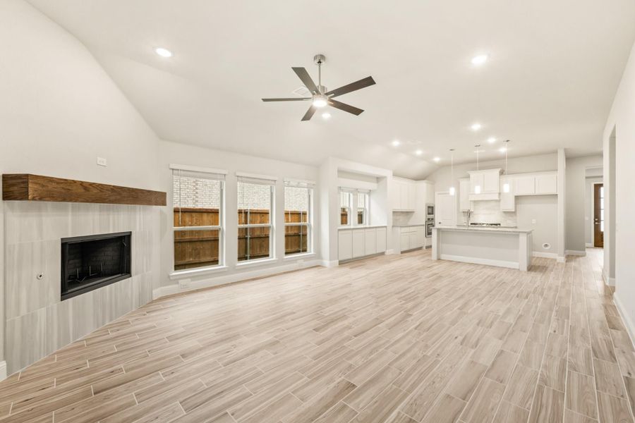 Open living room with tile fireplace and wood mantel, ceiling fan, and open kitchen with white cabinets and island Open living room with tile fireplace and wood mantel, ceiling fan, and open kitchen with white cabinets and island