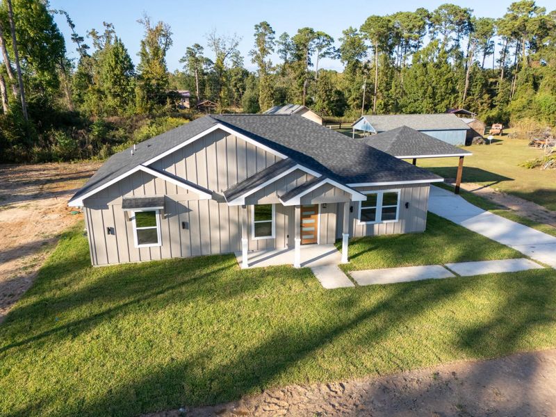 This charming home features a modern exterior with a gabled roof and large windows. It's set on a spacious, grassy lot with a concrete driveway. Surrounded by trees, it offers a serene, natural setting.