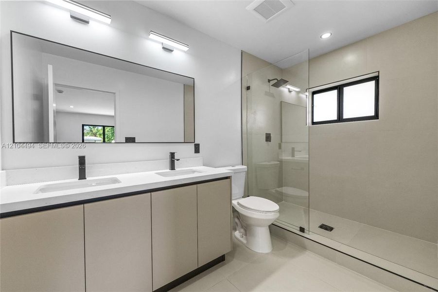 Dual-sink vanity with light-tone cabinetry and white countertop, large wall-mounted mirror, walk-in glass-enclosed shower, and matte black plumbing fixtures