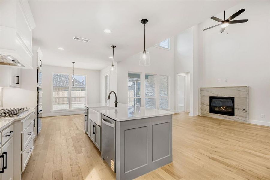 Open-concept living space with light wood-finish flooring, a contemporary kitchen island featuring a farmhouse sink, and a fireplace with a stone-finish surround