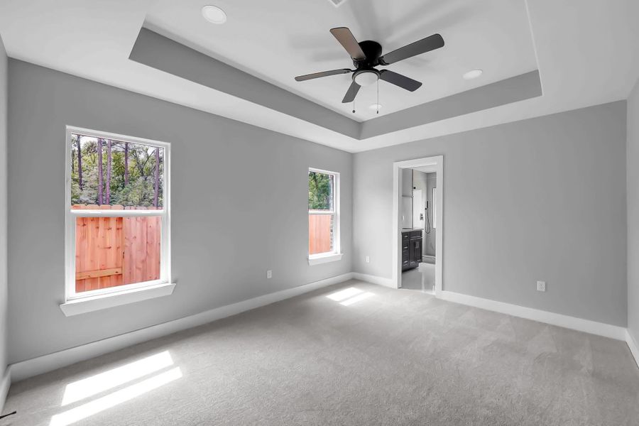 Bright and spacious bedroom with neutral gray walls and carpet. Features a tray ceiling with a modern ceiling fan and two large windows for natural light. There's an en-suite bathroom visible through an open doorway.