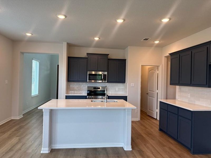 Contemporary kitchen featuring a center island with sink, dark blue cabinetry, light tile backsplash, and wood-finish flooring Contemporary kitchen featuring a center island with sink, dark blue cabinetry, light tile backsplash, and wood-finish flooring