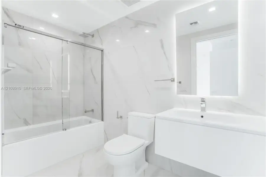 Bathroom featuring large-format porcelain tile, wall-mounted floating vanity, glass tub enclosure, and backlit vanity mirror