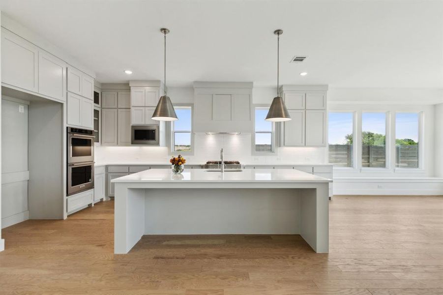 Kitchen featuring a large central island with a sink, white cabinetry, integrated stainless steel double ovens, and wood-finish flooring Kitchen featuring a large central island with a sink, white cabinetry, integrated stainless steel double ovens, and wood-finish flooring