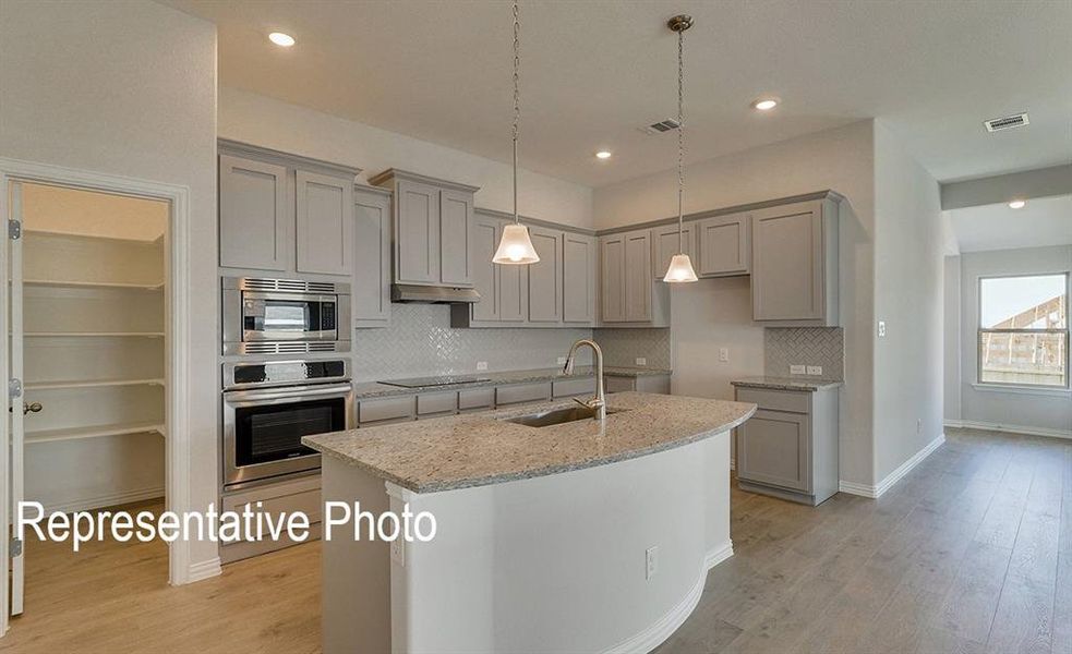 Modern kitchen featuring gray cabinetry, stainless steel appliances, a light-colored stone countertop island with an undermount sink, a white chevron-patterned tile backsplash, and wood-finish flooring