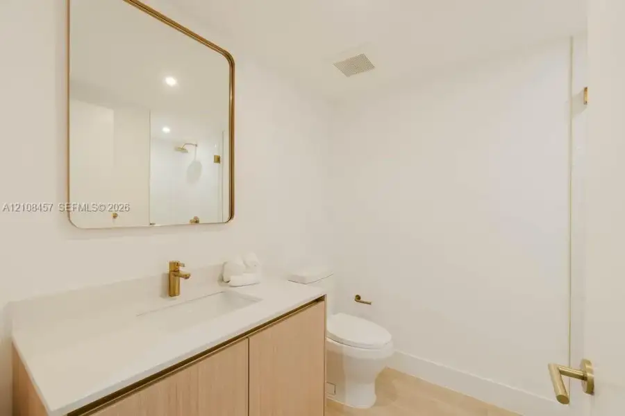 Modern vanity with wood-finish cabinetry, white countertop, integrated rectangular sink, brass-toned hardware, and a matching rounded-edge wall mirror Modern vanity with wood-finish cabinetry, white countertop, integrated rectangular sink, brass-toned hardware, and a matching rounded-edge wall mirror
