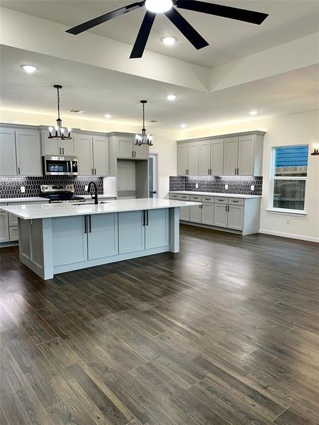 Kitchen with an island with sink, gray cabinetry, hanging light fixtures, stainless steel appliances, and dark wood-type flooring