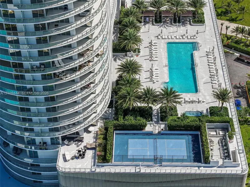 High-rise exterior featuring curved wraparound balconies, a swimming pool with paved deck, a tennis court, and manicured landscaping