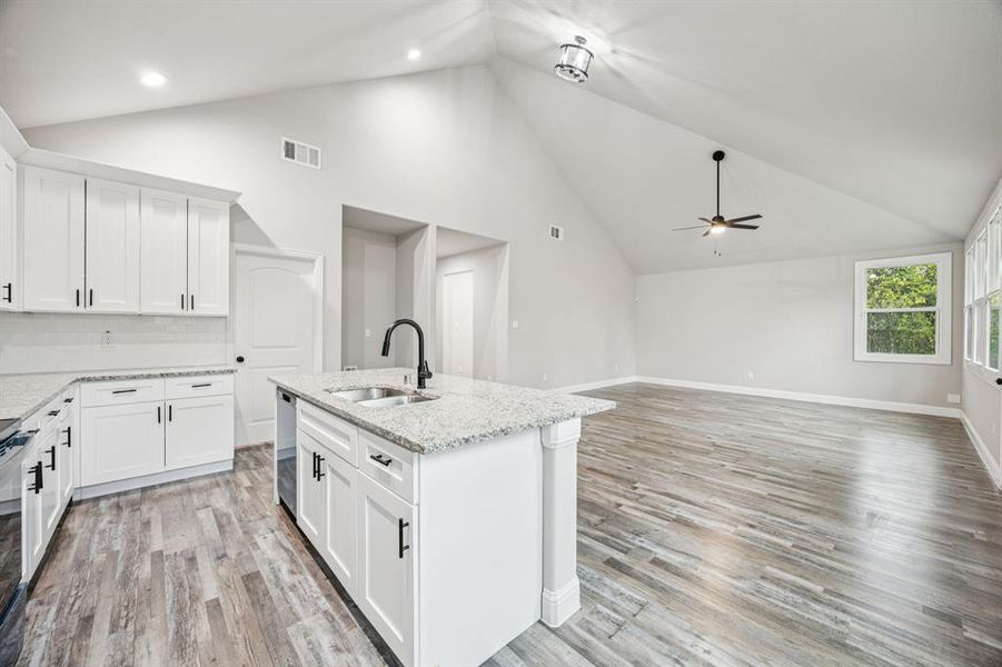 Kitchen featuring white cabinets, granite countertops, backsplash, a center island with sink, light wood-style floors, and light stone counters