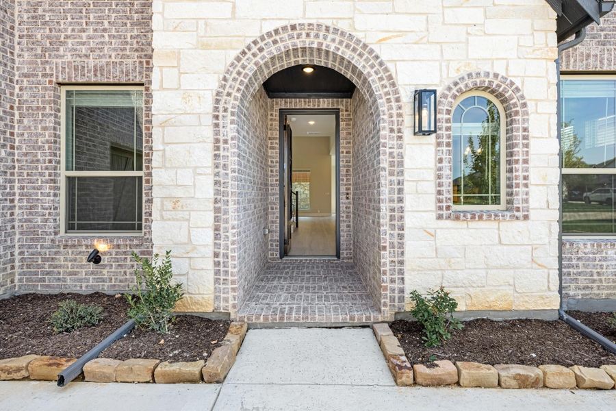 Home entrance with arched brick and stone surround, open front door, wall sconce, and concrete walkway with landscaping.