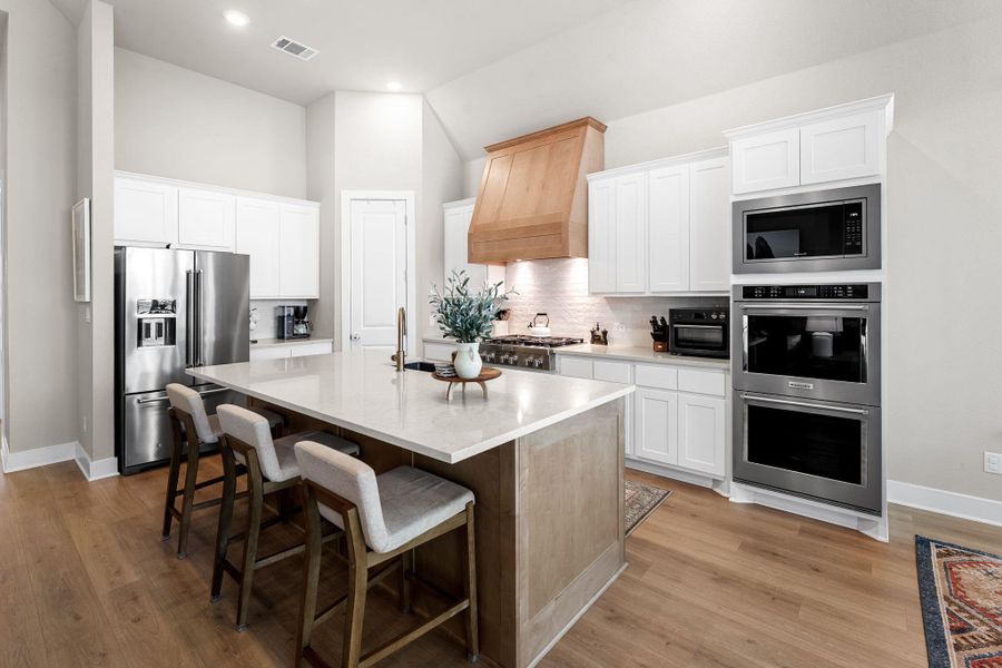 Kitchen featuring a large central island with a light-toned countertop, wood-finish flooring, white cabinetry, a natural wood range hood, and built-in stainless steel appliances
