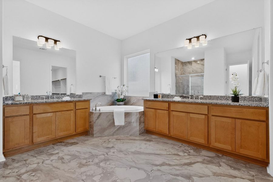 Spacious bathroom featuring an oval soaking tub with stone surround, dual vanities with light wood cabinetry and stone countertops, and large wall-mounted mirrors with integrated lighting