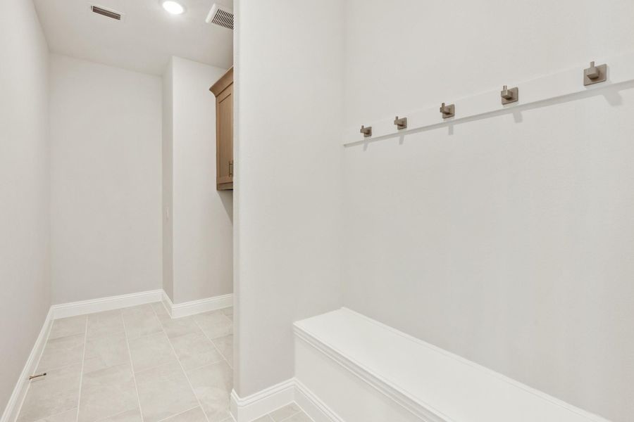 Mudroom with white built-in bench, row of metal hooks, and tile floor leading to storage cabinets