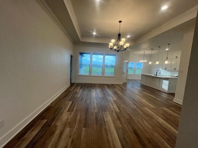 This spacious, open-concept living area features elegant medium gray/wood water proof plank flooring, a modern chandelier, and abundant natural light from large windows. The adjacent kitchen boasts stylish pendant lighting and a sleek island perfect for entertaining.