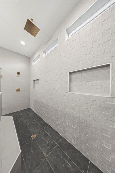 Spacious shower with a sloped ceiling, rectangular rain shower head, textured white wall tile, dark stone-finish floor tile, and multiple recessed wall niches