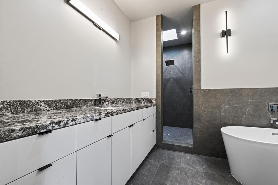 The primary bath delivers a spa-like experience: a freestanding soaking tub, an oversized walk-in shower finished in large-format tile with rain showerhead, and a skylight above that floods the space with natural light. (Mirrors to be installed)