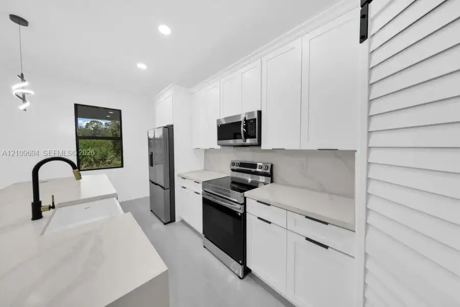 Kitchen featuring white shaker-style cabinetry, quartz countertops with integrated backsplash, stainless steel appliances, a farmhouse-style sink with a matte black faucet, and a sliding louvered barn door