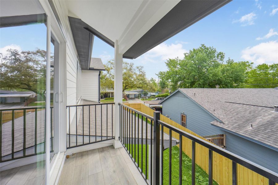 This photo features a cozy balcony with sleek black railing and wood-look flooring, offering a view of neighboring rooftops and lush greenery. It's an inviting outdoor space for relaxation.