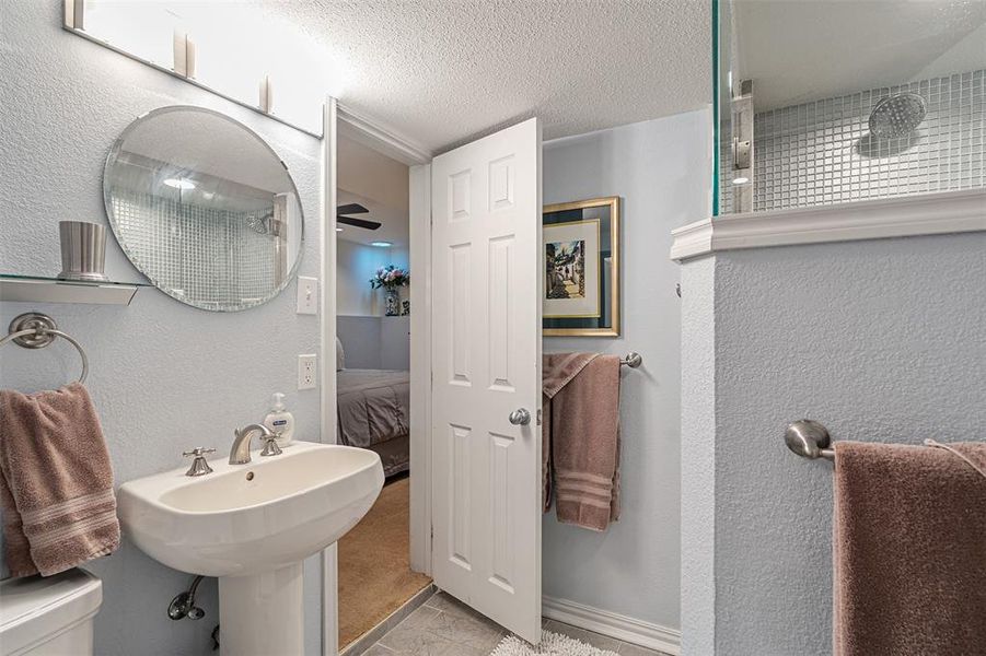 Bathroom featuring a pedestal sink with a brushed nickel faucet, a round wall-mounted mirror, and a glass-enclosed shower with small square tile