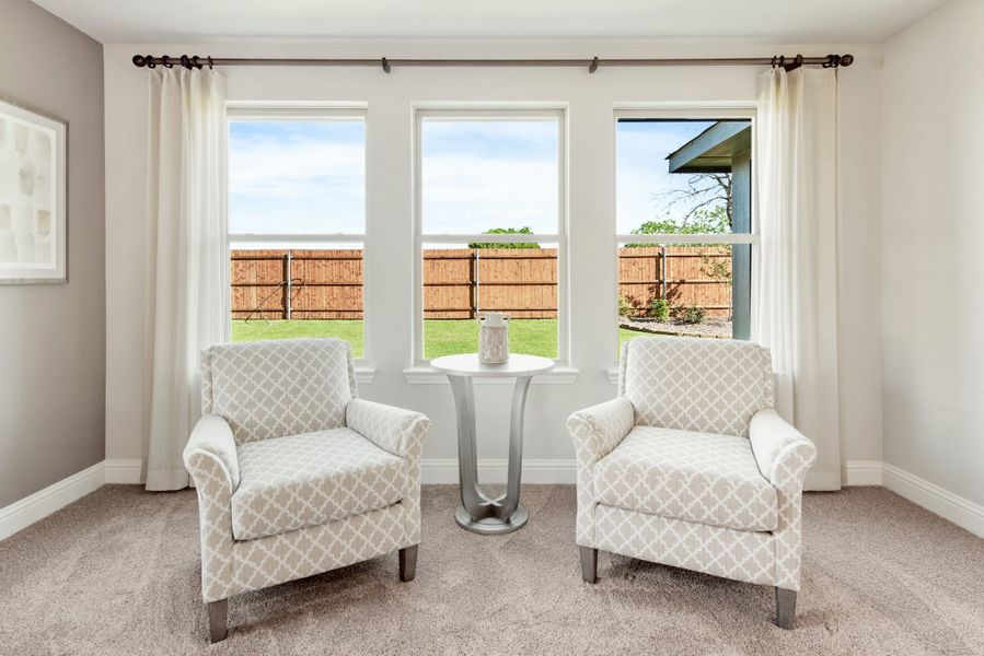 Sitting area with two patterned armchairs, small round table, and triple windows overlooking fenced backyard