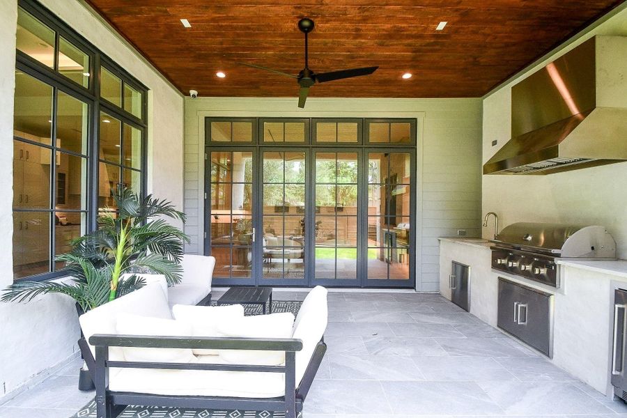 This photo showcases a stylish outdoor living area with a modern kitchen setup, featuring a built-in grill and stainless steel appliances. The space is complemented by comfortable seating, a wooden ceiling, and large glass doors that open to the garden, creating a seamless indoor-outdoor flow.