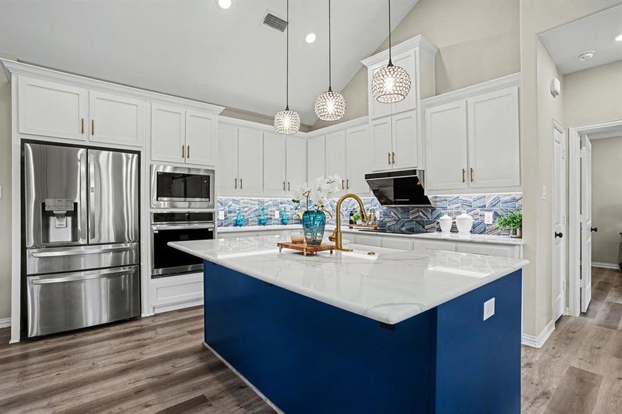 Two tone kitchen with stainless steel appliances, an island with farm sink, light stone counters, dark wood-style floors, and hanging light fixtures