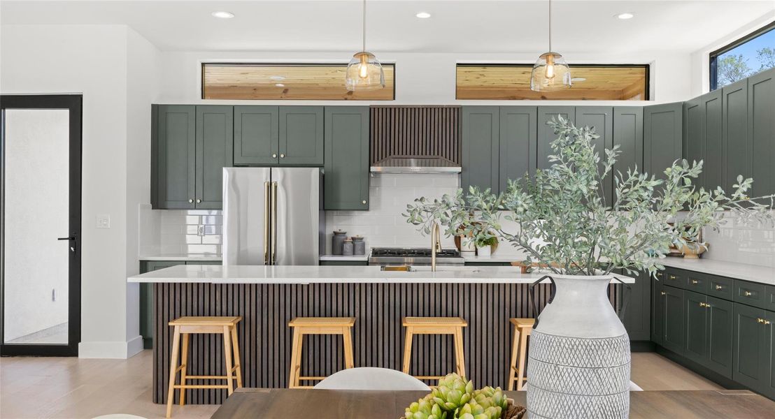 Kitchen featuring green cabinetry, a kitchen island with sink, high quality fridge, a kitchen bar, and decorative light fixtures