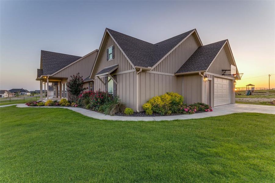 Property exterior at dusk with a shingled roof, an attached garage, a lawn, board and batten siding, and driveway