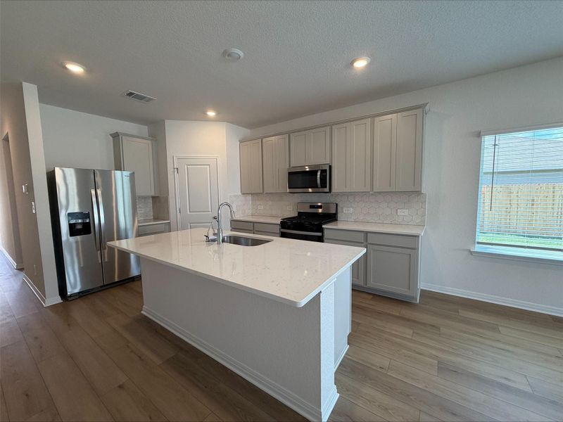 Kitchen featuring appliances with stainless steel finishes, light stone counters, tasteful backsplash, gray cabinetry, and a center island with sink Kitchen featuring appliances with stainless steel finishes, light stone counters, tasteful backsplash, gray cabinetry, and a center island with sink