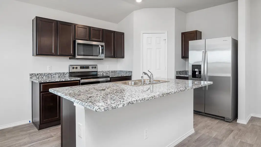 Kitchen with stainless steel appliances, dark wood finish cabinetry, light stone countertops, an island with sink, and light wood finished floors