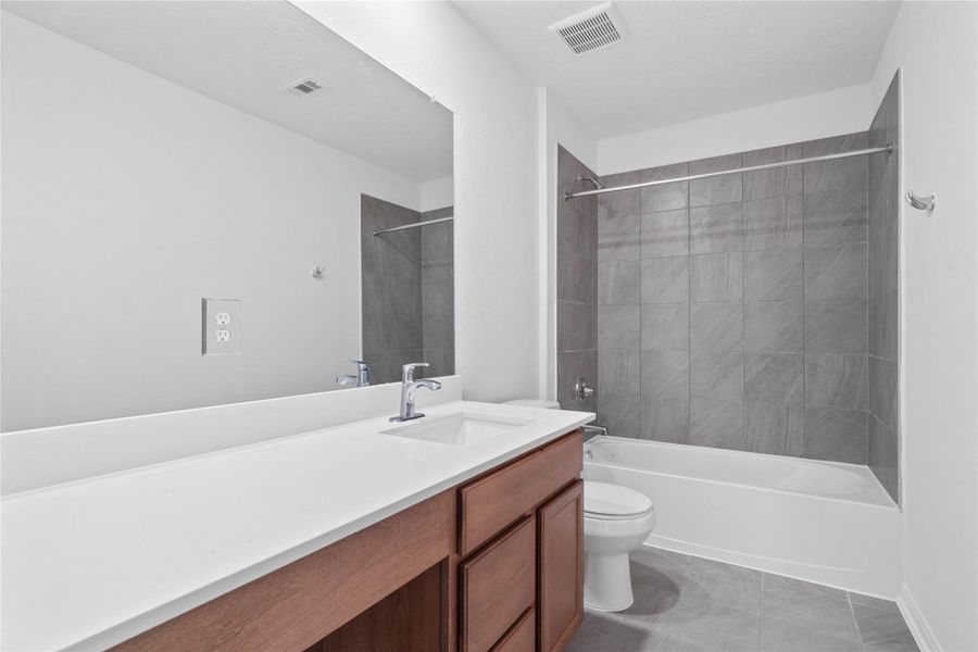The secondary bathroom offers tile flooring, a bath/shower combination with a stylish tile surround, rich stained wood cabinetry with added knee space and elegant light countertops, a mirror, and sleek fixtures with modern finishes.