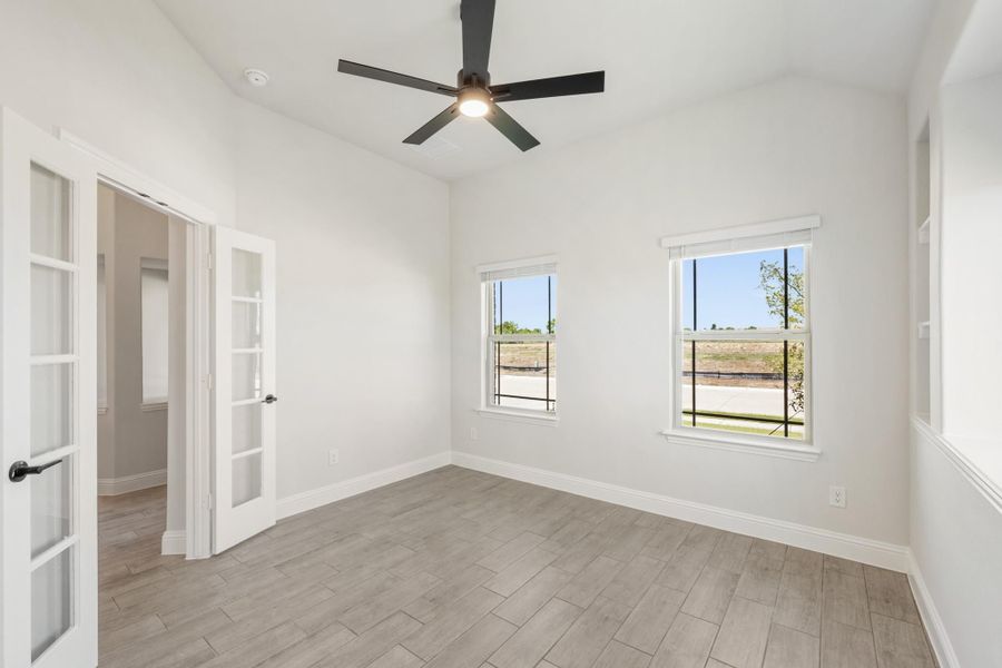 Empty room with wood-look tile floors, ceiling fan, two windows, and French doors leading to hallway Empty room with wood-look tile floors, ceiling fan, two windows, and French doors leading to hallway