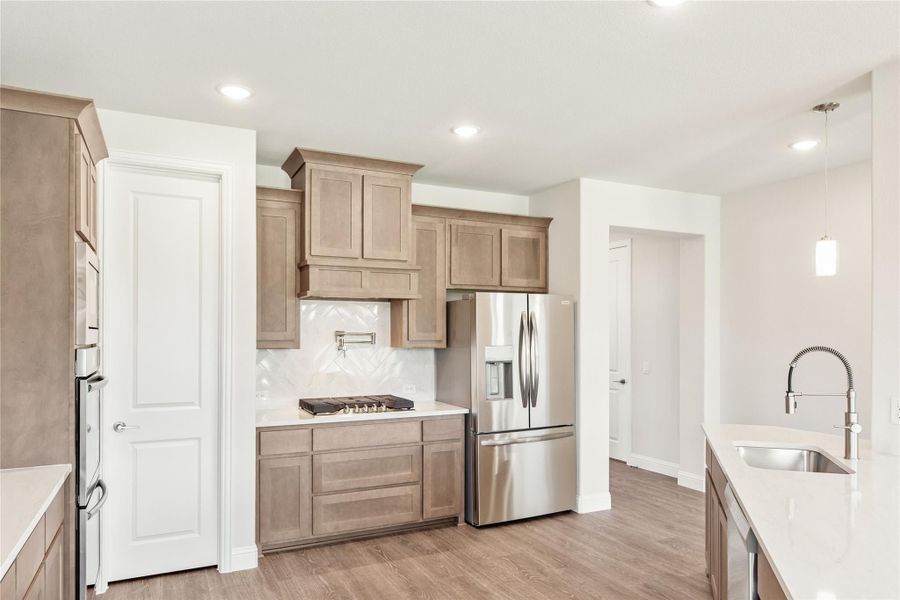 Kitchen with tan wood cabinets, stainless steel refrigerator, gas cooktop, herringbone backsplash, and hardwood floors