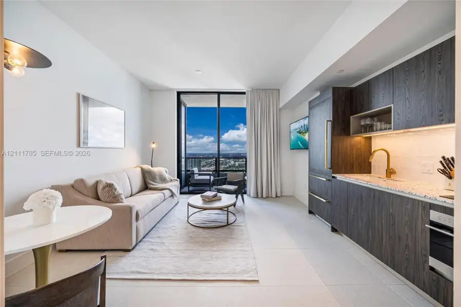 Kitchen featuring dark wood-finish cabinetry, under-cabinet lighting, stone-surface countertops, and gold-toned faucet hardware, floor-to-ceiling glass doors opening to a balcony with an expansive urban backdrop