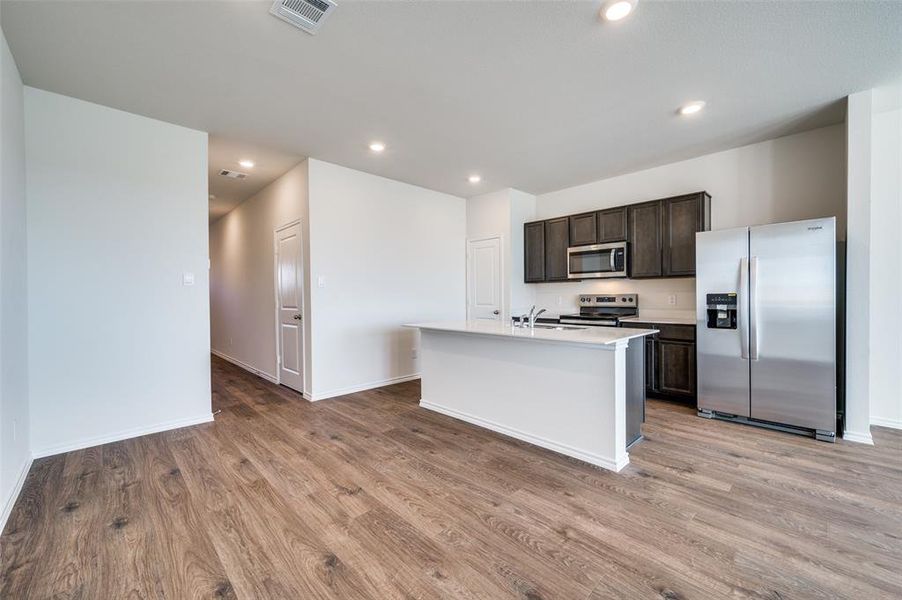 Kitchen with light wood-type flooring, stainless steel appliances, dark brown cabinetry, and a center island with sink