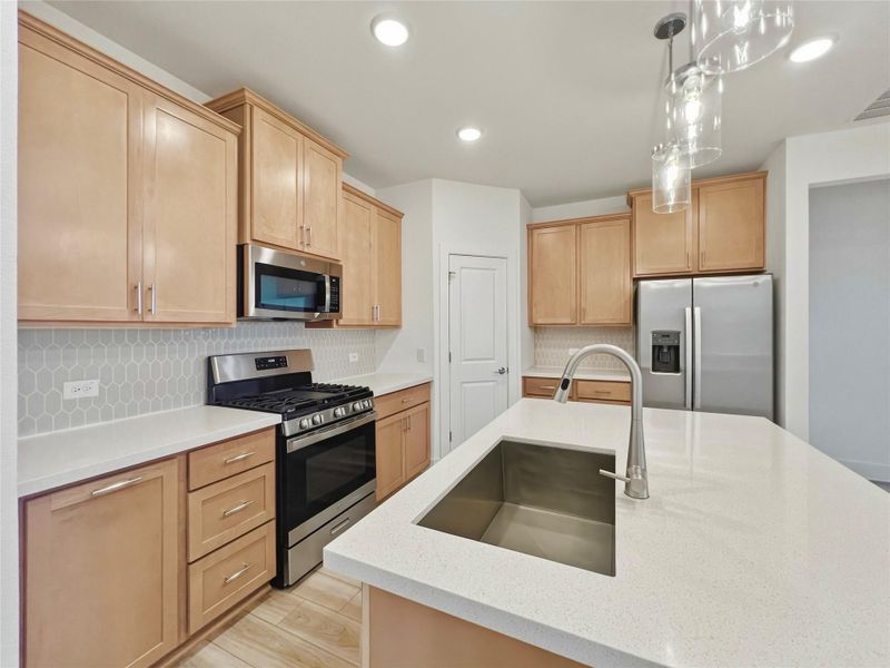 Kitchen with stainless steel appliances, hanging light fixtures, decorative backsplash, light stone counters, and a kitchen island with sink