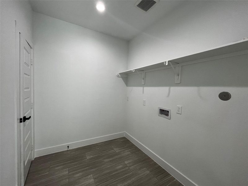 This photo shows a small, well-lit laundry room with white walls, a tiled floor, and a shelf. It has connections for a washer and dryer and a door leading to a closet that can be used for storage to the left.