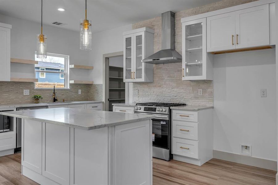 Kitchen with white cabinetry, wall chimney exhaust hood, appliances with stainless steel finishes, glass insert cabinets, and pendant lighting Kitchen with white cabinetry, wall chimney exhaust hood, appliances with stainless steel finishes, glass insert cabinets, and pendant lighting