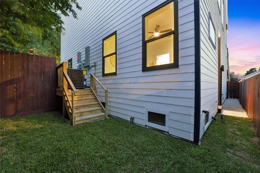 Private, fenced side yard featuring lush grass, wood steps leading to an elevated deck, modern exterior siding, and evening lighting—offering a functional, low-maintenance outdoor space with easy home access.