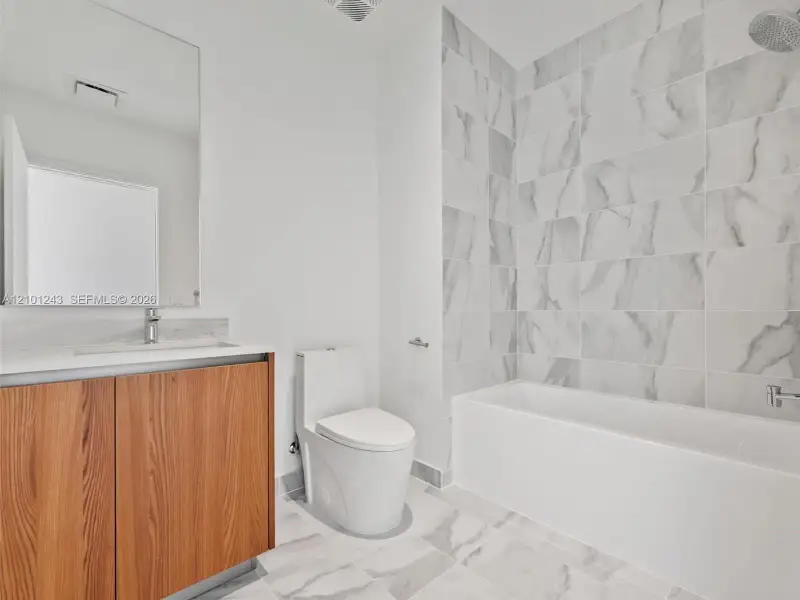 Bathroom featuring a wood-finish vanity with a stone-surface countertop, floor-to-ceiling tile shower surround, and large-format tile flooring Bathroom featuring a wood-finish vanity with a stone-surface countertop, floor-to-ceiling tile shower surround, and large-format tile flooring